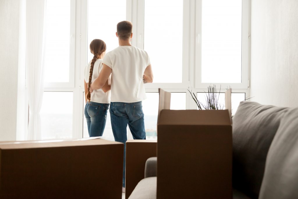 Millennial couple stand near window looking far away after moving in to new home with cardboard boxes, husband and wife plan future together, hugging seeing new perspectives. New beginning concept